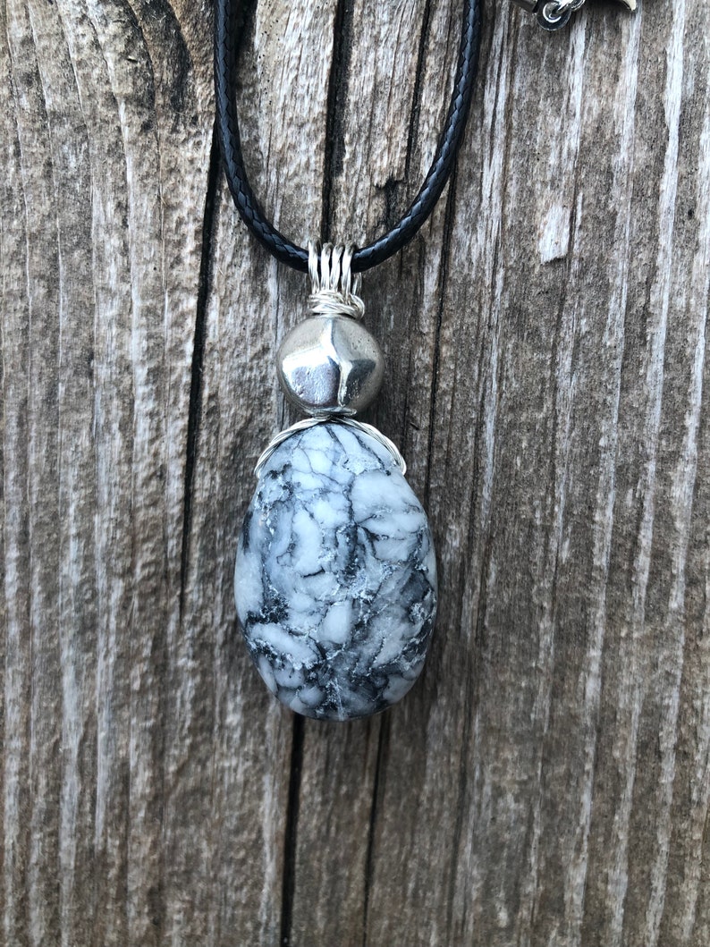 May include: A silver pendant necklace with a black cord. The pendant features a round silver bead and a larger, oval-shaped, white and grey stone with a marbled pattern.