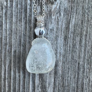 Op de afbeelding: Een zilveren ketting met een heldere glazen hanger die is gewikkeld in zilverdraad en een zilveren kraal.