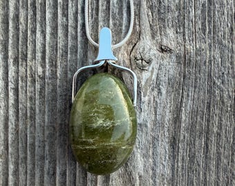 Op de afbeelding: Een zilveren hangerketting met een grote, ovale groene steen. De gepolijste steen hangt aan een zilveren ketting. De hanger staat tegen een verweerde houten achtergrond. De steen is ongeveer 3 cm lang.