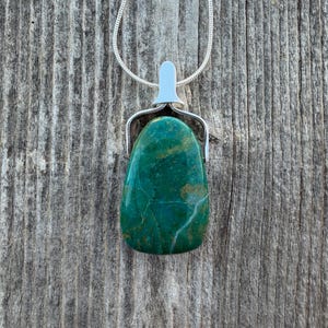 May include: A polished, teardrop-shaped pendant necklace featuring a vibrant green stone with natural veining. The stone is held in a silver-colored metal setting and hangs from a delicate silver chain. The necklace is displayed on a weathered wooden surface.