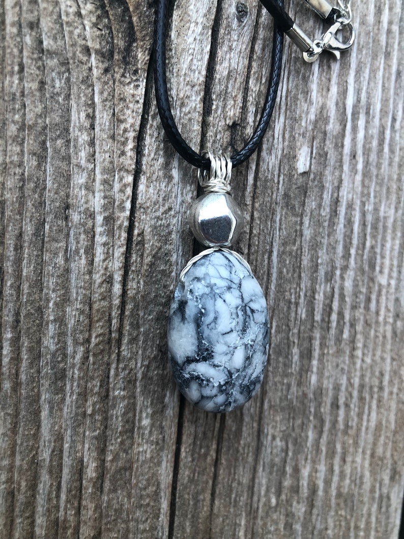 May include: A black leather cord necklace with a silver pendant featuring a white and grey stone. The stone has a unique marbled pattern.