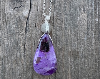 May include: A silver chain necklace with a purple and black teardrop-shaped pendant. The pendant is made of a natural stone with a unique pattern.