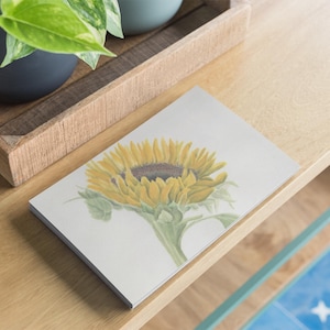 Könnte beinhalten: Ein rechteckiges Notizbuch mit einer detaillierten Aquarellillustration einer einzelnen gelben Sonnenblume mit dunkelbrauner Mitte und grünen Blättern auf einem weißen Einband. Das Notizbuch liegt flach auf einer hellen Holzoberfläche.