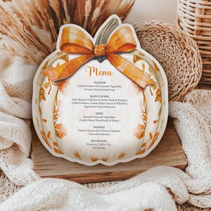 May include: A pumpkin-shaped menu card with an orange and yellow plaid bow. The menu features a fall-themed menu with starters, main courses, sides, desserts, and beverages. The card is on a wooden board with a white knitted blanket.