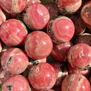 K&ouml;nnte beinhalten: Eine Sammlung von rosa und wei&szlig;en Rhodochrosit-Kugeln. Die Kugeln sind glatt und poliert und haben verschiedene Muster und Texturen.
