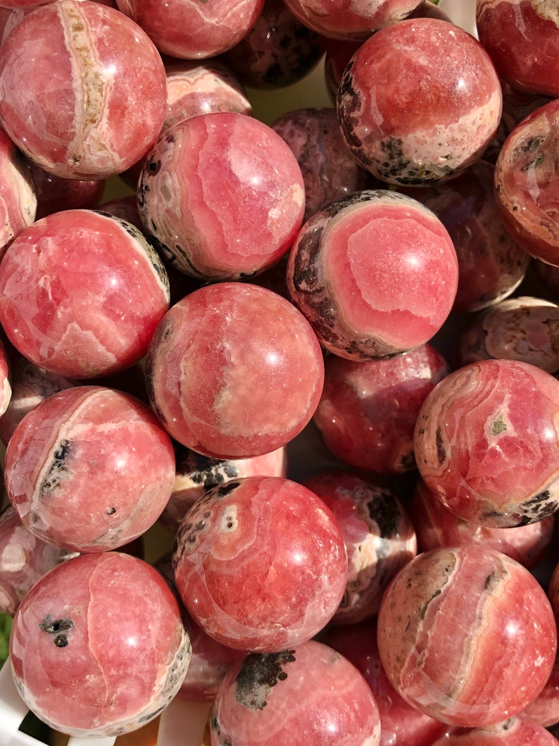 K&ouml;nnte beinhalten: Eine Sammlung von rosa und wei&szlig;en Rhodochrosit-Kugeln. Die Kugeln sind glatt und poliert und haben verschiedene Muster und Texturen.