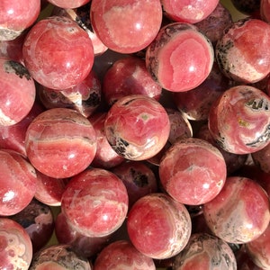 K&ouml;nnte beinhalten: Eine Sammlung von rosa und schwarzen Rhodochrosit-Kugeln. Die Kugeln sind glatt und poliert und haben verschiedene Muster und Texturen.