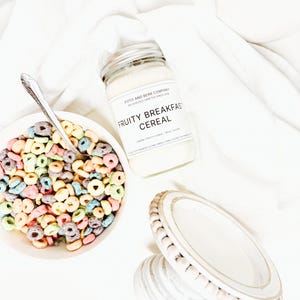 May include: A glass jar candle labeled Fruity Breakfast Cereal with a white wax fill, positioned next to a white bowl filled with colorful ring-shaped cereal and a silver spoon, and a small white wooden pedestal stand with beaded trim.