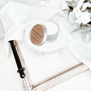 May include: A small, silver candle tin with a brown label that reads "Toasted Pistachio and Salted Caramel" sits on a white plate. The candle is partially open, revealing the white wax inside. A distressed white picture frame is in the foreground.