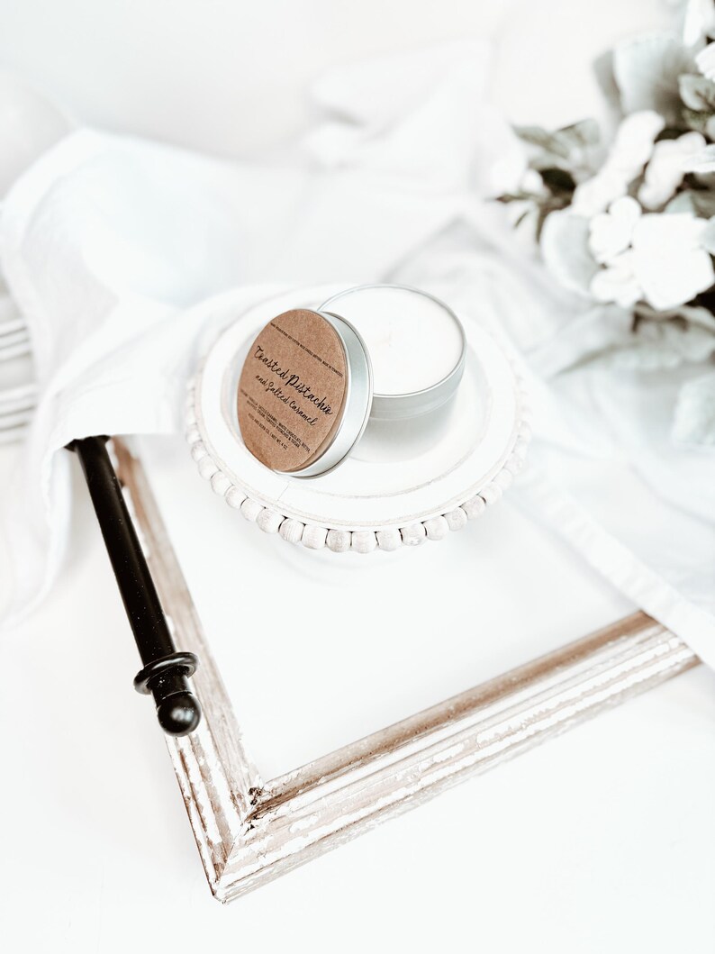 May include: A small, silver candle tin with a brown label that reads "Toasted Pistachio and Salted Caramel" sits on a white plate. The candle is partially open, revealing the white wax inside. A distressed white picture frame is in the foreground.