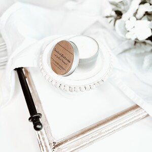 May include: A small, silver candle tin with a brown label that reads "Toasted Pistachio and Salted Caramel" sits on a white plate. The plate is on a white surface with a distressed white picture frame and white fabric.