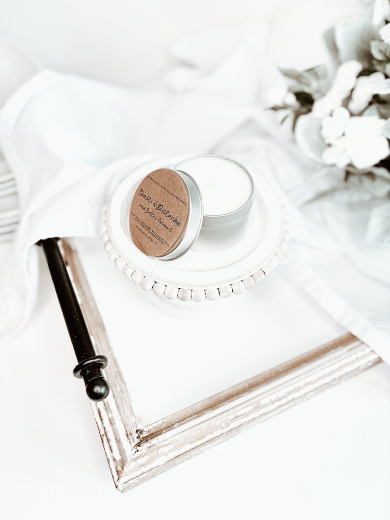 May include: A small, silver candle tin with a brown label that reads "Toasted Pistachio and Salted Caramel" sits on a white plate. The plate is on a white surface with a distressed white picture frame and white fabric.
