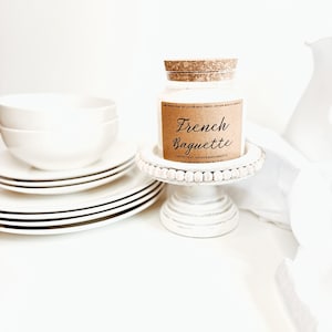May include: A white candle in a glass jar with a cork lid, labeled "French Baguette." The candle sits on a white, beaded cake stand next to a stack of white plates with black trim and two white bowls. The background is white.