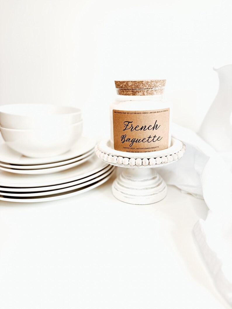 May include: A white candle in a glass jar with a cork lid, labeled "French Baguette." The candle sits on a white, beaded cake stand next to a stack of white plates with black trim and two white bowls. The background is white.