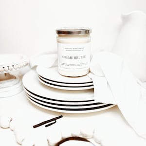 May include: A glass jar candle with a silver lid and a white label reading Creme Brulee, Poto and Burr Company, and scent notes including caramelized sugar crust, torched sugar, creamy custard, coconut, rum, maple, and sugar, resting on white plates.