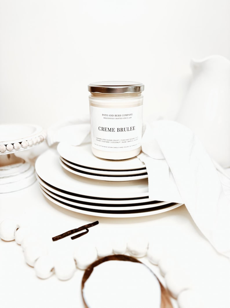 May include: A glass jar candle with a silver lid and a white label reading Creme Brulee, Poto and Burr Company, and scent notes including caramelized sugar crust, torched sugar, creamy custard, coconut, rum, maple, and sugar, resting on white plates.