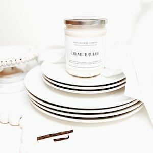 May include: A glass jar candle with a silver lid, labeled Poto and Berr Company Creme Brulee, resting on a stack of white plates. The label lists notes of caramelized sugar crust, torched sugar, nut, creamy custard, coconut, rum, maple, and sugar.