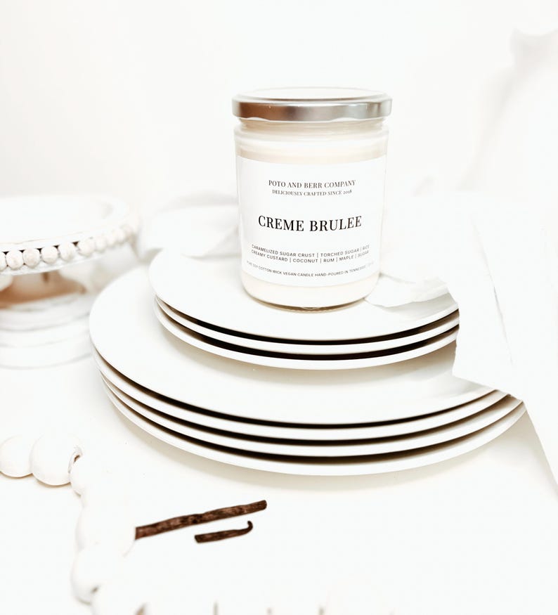 May include: A glass jar candle with a silver lid, labeled Poto and Berr Company Creme Brulee, resting on a stack of white plates. The label lists notes of caramelized sugar crust, torched sugar, nut, creamy custard, coconut, rum, maple, and sugar.