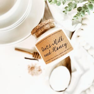 May include: A glass jar candle with a cork lid, labeled "Oats Milk and Honey." The label lists ingredients and the net weight of 22 oz. The candle is surrounded by white plates, a wooden honey dipper, and greenery.