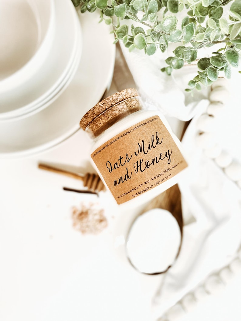 May include: A glass jar candle with a cork lid, labeled "Oats Milk and Honey." The label lists ingredients and the net weight of 22 oz. The candle is surrounded by white plates, a wooden honey dipper, and greenery.