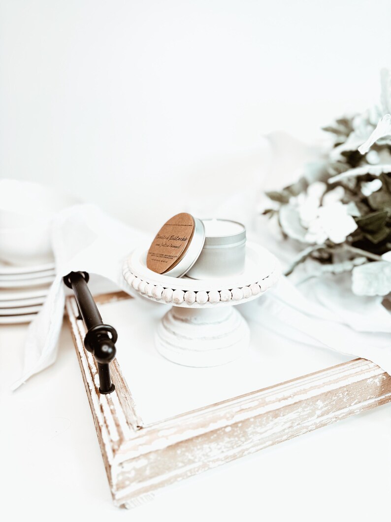 May include: A small, round, silver candle tin with a wooden lid sits atop a white, beaded cake stand. The stand is on a white tray with a distressed, light-colored wooden frame and a black handle. A white cloth and decorative flowers are in the background.
