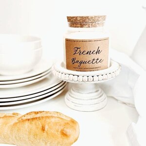 May include: A white jar candle with a cork lid sits on a white beaded cake stand. The candle has a brown label that reads "French Baguette." A loaf of bread and a stack of white plates are also in the image.