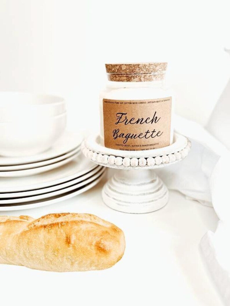 May include: A white jar candle with a cork lid sits on a white beaded cake stand. The candle has a brown label that reads "French Baguette." A loaf of bread and a stack of white plates are also in the image.