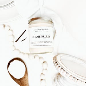 May include: A clear glass candle jar with a silver lid and a white label reading Poto and Berr Company, Creme Brulee, and a list of scent notes including caramelized sugar crust, torched sugar, creamy custard, coconut, rum, maple, and sugar.
