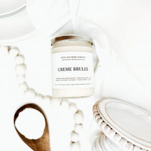 May include: A clear glass candle jar with a silver lid and a white label featuring the text Poto and Berr Company, Creme Brulee, and a list of scent notes including caramelized sugar crust, torched sugar, creamy custard, coconut, rum, and maple.