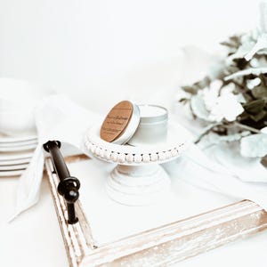 May include: A small, silver candle tin with a wooden lid sits atop a white, beaded cake stand. The stand is on a white tray with a distressed, light-colored wooden frame and a black handle. A white cloth and decorative flowers are in the background.