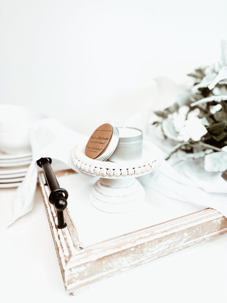 May include: A small, silver candle tin with a wooden lid sits atop a white, beaded cake stand. The stand is on a white tray with a distressed, light-colored wooden frame and a black handle. A white cloth and decorative flowers are in the background.