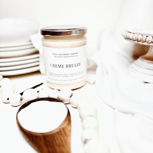 May include: A clear glass candle jar with a silver lid and a white label featuring the text Poto and Berr Company, Deliciously Crafted Since 2018, and Creme Brulee. The candle contains off-white wax and is positioned near a wooden spoon.