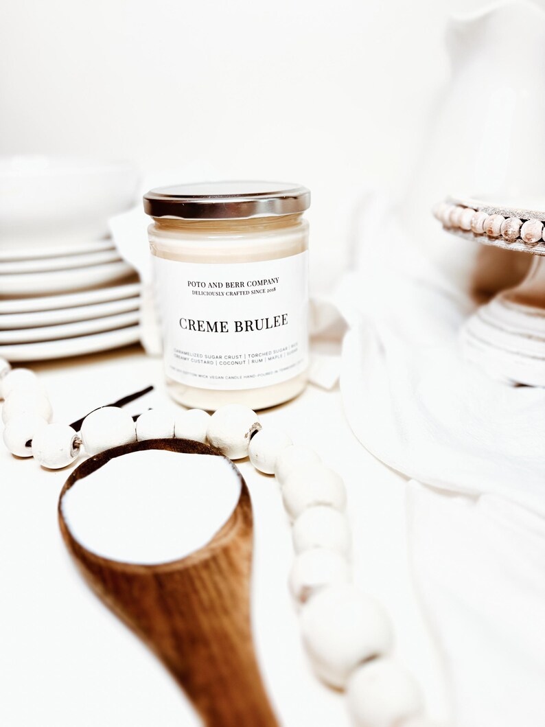 May include: A clear glass candle jar with a silver lid and a white label featuring the text Poto and Berr Company, Deliciously Crafted Since 2018, and Creme Brulee. The candle contains off-white wax and is positioned near a wooden spoon.