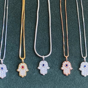 May include: Six silver, gold, and rose gold necklaces with hamsa hand pendants. Each pendant is adorned with clear cubic zirconia stones and a blue or red gemstone in the center.