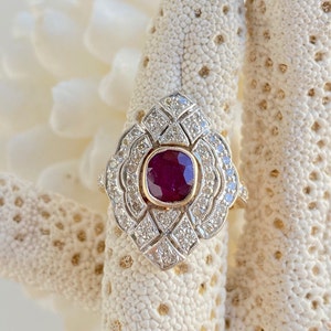 May include: A vintage-style ring with a large, deep red ruby surrounded by a halo of smaller diamonds. The ring is set in yellow gold and has a delicate, intricate design.
