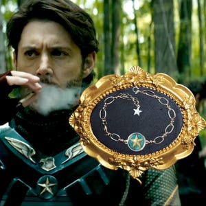 Puede incluir: Pulsera plateada con un dije de estrella y un colgante circular con una estrella dorada sobre fondo verde. La pulsera se exhibe sobre una superficie negra dentro de un marco dorado ornamentado.