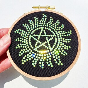 May include: A black fabric hoop with a green and gold sparkly star design. The star is surrounded by a circle of smaller green and gold sparkly dots.