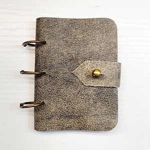 May include: A small gray leather bound journal with three metal rings and a flap closure with a brass button.