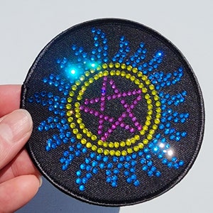 May include: A round black patch with a five-pointed star in the center. The star is made of pink and purple rhinestones and is surrounded by a yellow circle of rhinestones. The yellow circle is surrounded by a blue circle of rhinestones.