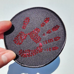 May include: A black fabric patch with a red rhinestone design. The design is abstract and resembles a stylized skull.