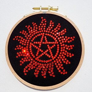 May include: A red and black embroidered hoop art piece featuring a pentagram with a sunburst design. The pentagram is made of red rhinestones and the sunburst is made of red beads.