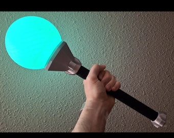 May include: A glowing teal orb atop a black and silver staff. The orb emits a bright, even light. The staff is held in a hand, suggesting a prop or decorative item. The design is futuristic and fantasy-themed.