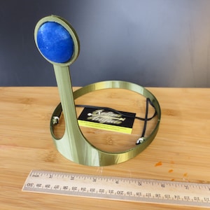 May include: A green plastic headband with a blue round gem on top. The headband is designed to be worn on the head and has a black elastic band for a secure fit.
