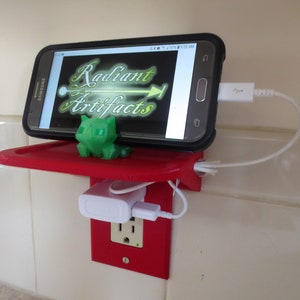 May include: A red 3D printed wall mount with a shelf for a phone and a USB charging port. The phone is charging with a white cable. The wall mount is attached to a standard electrical outlet.