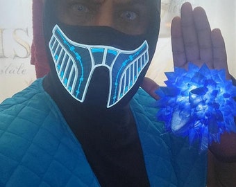 May include: A person wearing a black hood and mask with a blue and white illuminated design. The person is also wearing a blue quilted jacket and holding a glowing blue object.