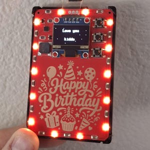 May include: A rectangular red electronic circuit board with a small digital screen displaying the text Love you kiddo. The board features a Happy Birthday graphic with party icons and is illuminated by a border of bright red LED lights.