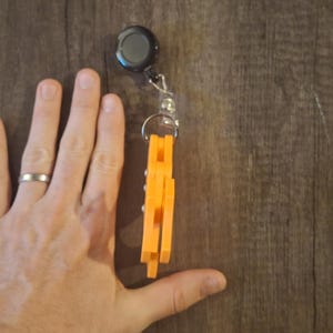 May include: An orange retractable badge holder with a black top and a silver chain. A small, pink and white card holder with a cartoon design is also visible. The items are on a wooden surface.