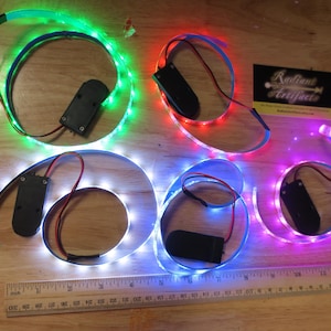 Puede incluir: Cinco tiras de luz LED de diferentes colores con paquetes de baterías. Las tiras son verde, roja, azul, blanca y rosa. Las tiras están enrolladas y los paquetes de baterías son visibles.
