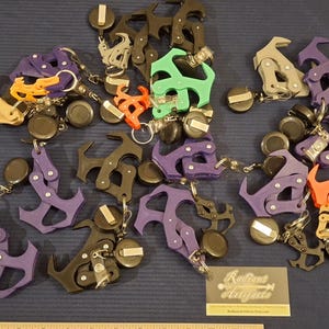 May include: A collection of colorful keychains in various shapes, including anchor and abstract designs. The keychains are in shades of orange, purple, green, and black, with silver-tone metal accents and black circular attachments. A business card is visible.