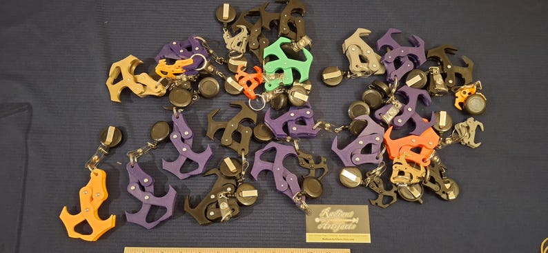 May include: A collection of colorful keychains in various shapes, including anchor and abstract designs. The keychains are in shades of orange, purple, green, and black, with silver-tone metal accents and black circular attachments. A business card is visible.